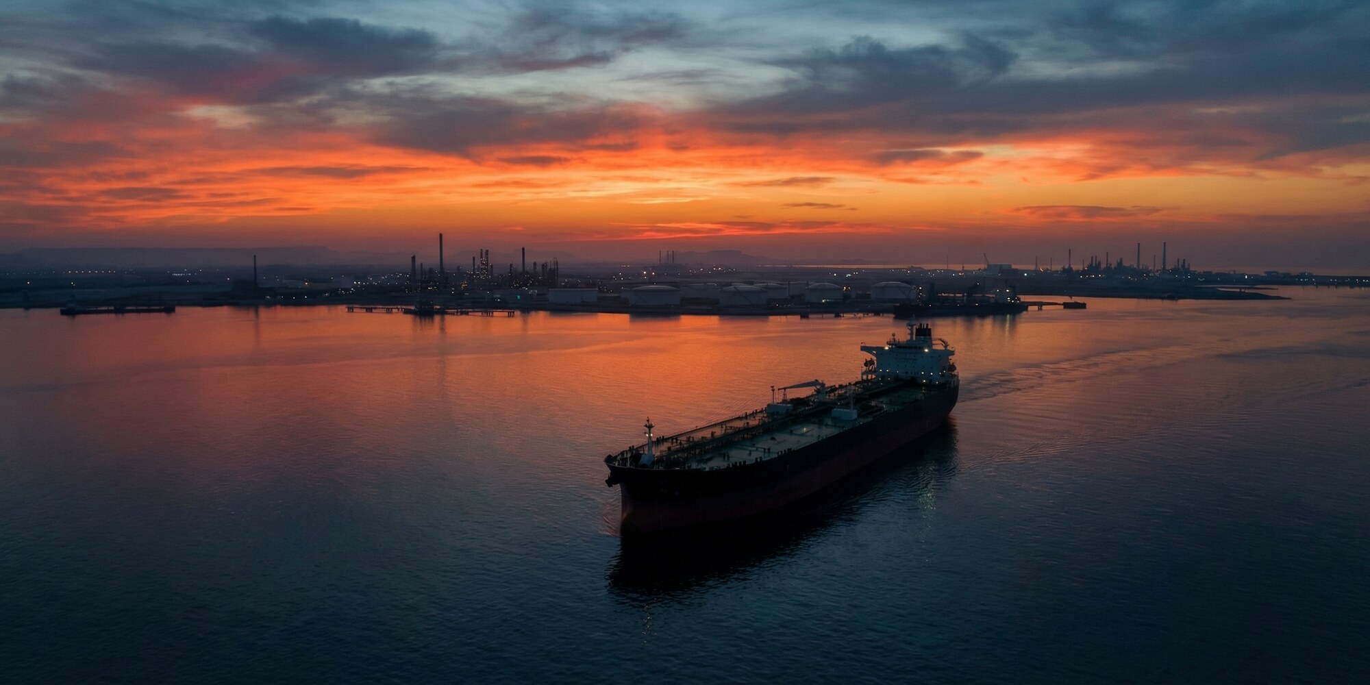 Oil supertanker navigating the Strait of Hormuz at dusk with refinery infrastructure on the horizon
